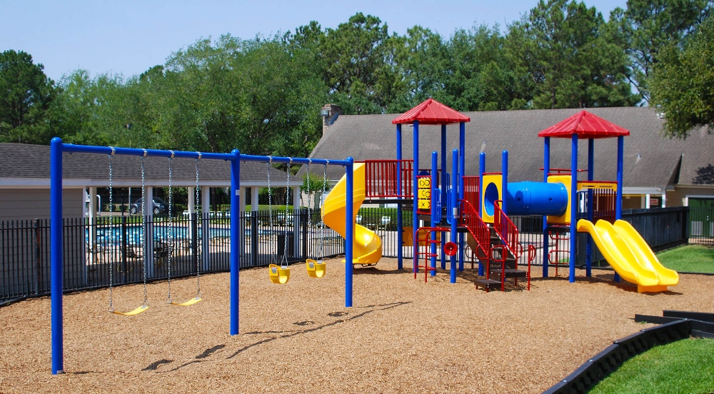Colorful commercial playground with blue swing set, yellow slides, and climbing structures installed over engineered wood fiber safety surfacing near a residential community pool area.